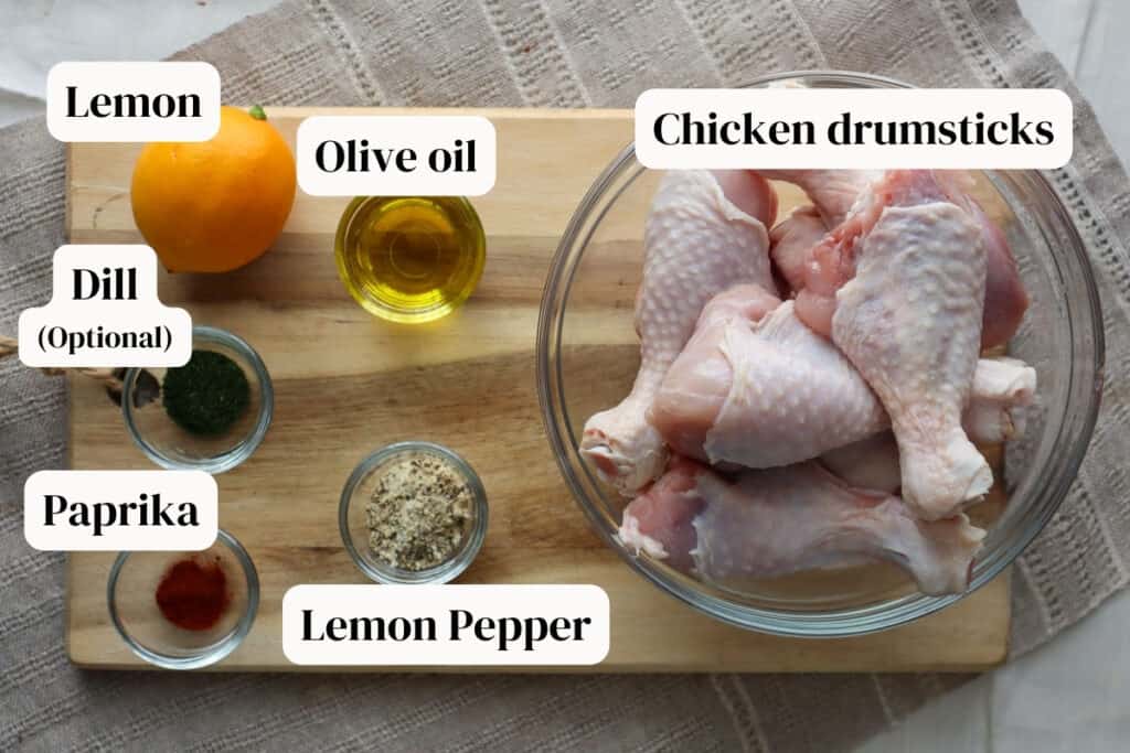 Chicken drumsticks in a glass bowl, olive oil, lemon pepper seasoning, dried dill, paprika in small glass bowls and a lemon, all on a wooden board.