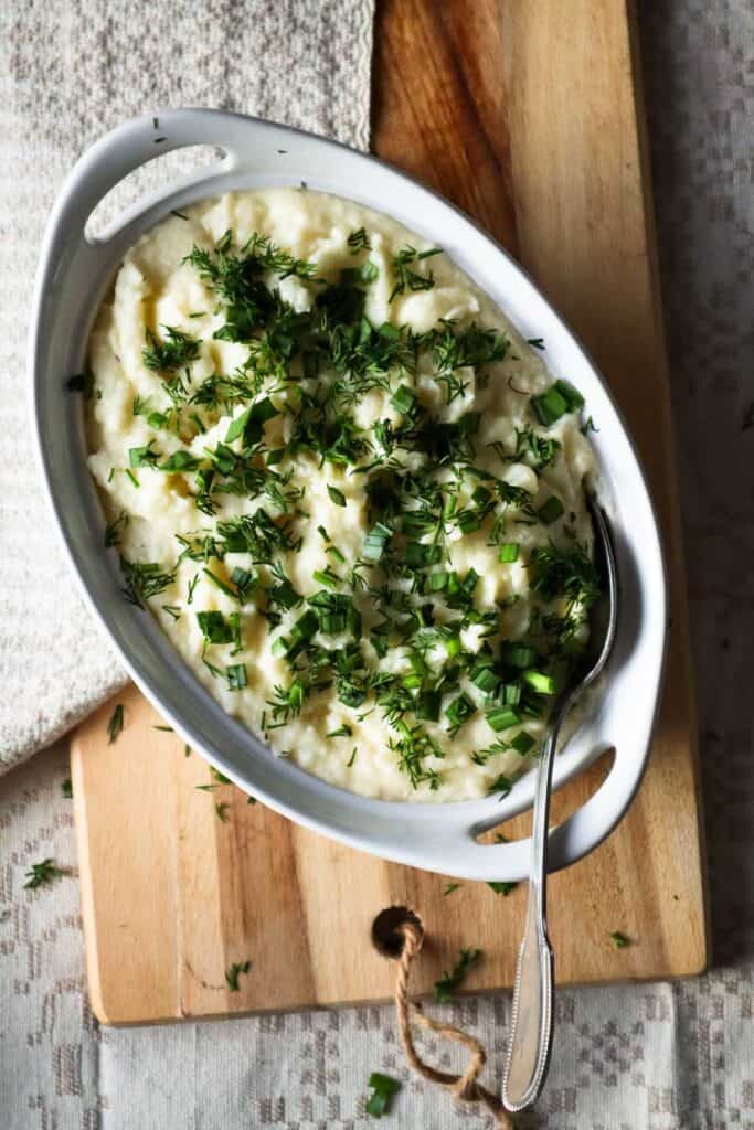Cottage cheese mashed potatoes with fresh herbs in white dish on wooden board.