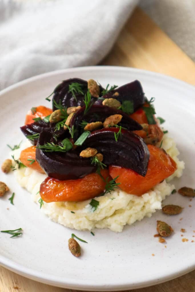 Mashed potatoes cottage cheese with roast beets and carrots topped with maple sunflower seeds, all on a beige plate.