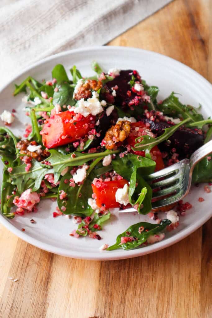Fall quinoa salad with roasted beets and carrots, baby spinach, arugula, feta and walnuts on a small beige plate and a forkful of salad.