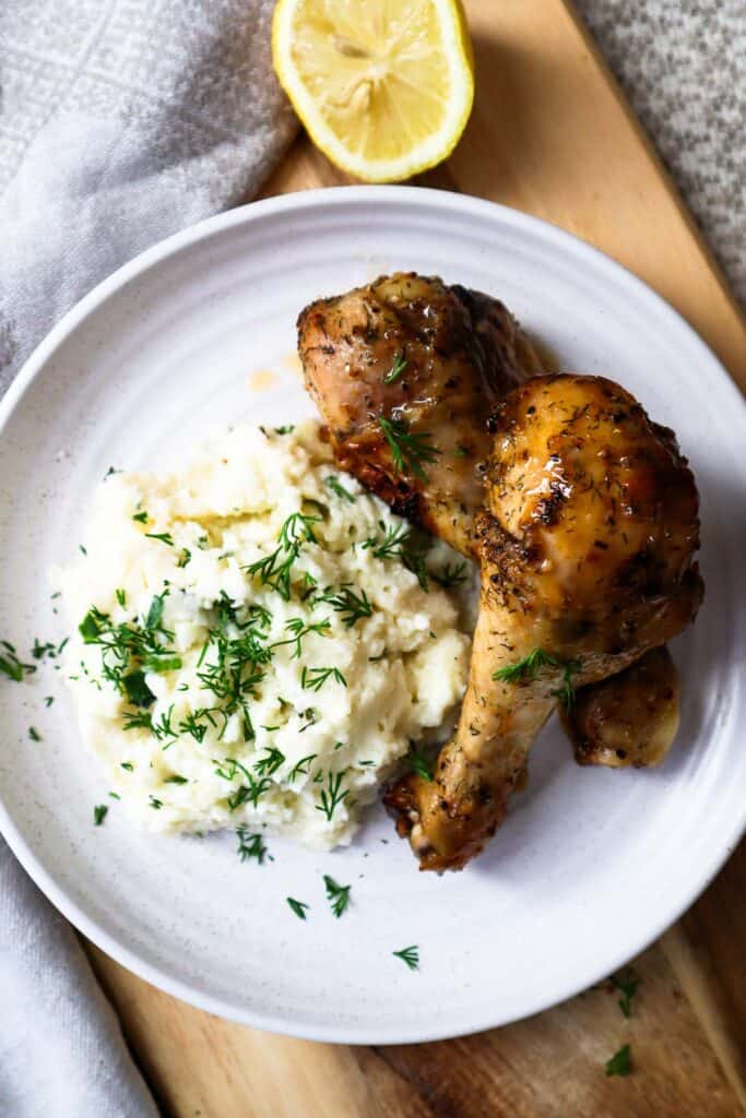Two lemon pepper chicken drumsticks on a beige plate with cotage cheese mashed potatoes and a half of a lemon.