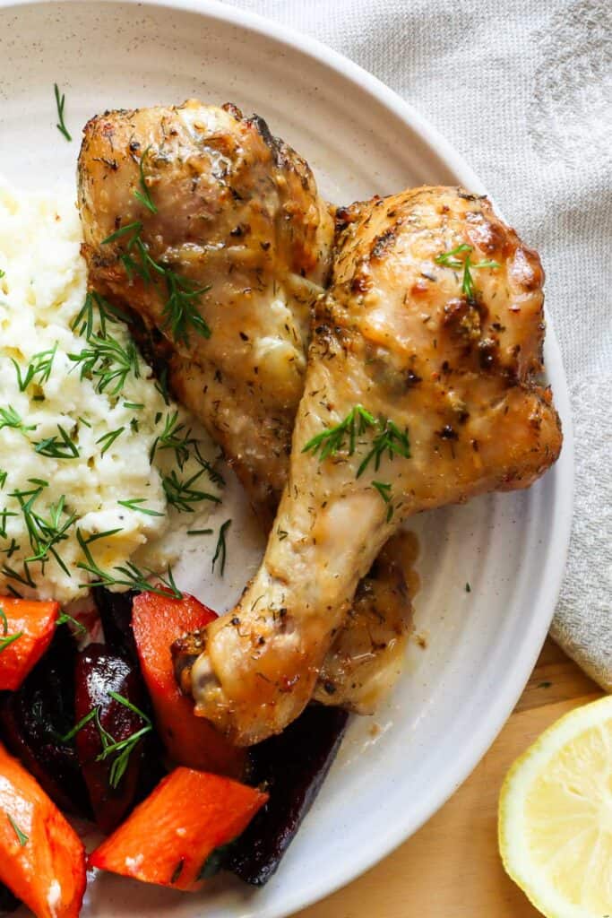 Two baked chicken drumsticks on beige plate with mashed potatoes and beets and carrots with fresh dill.