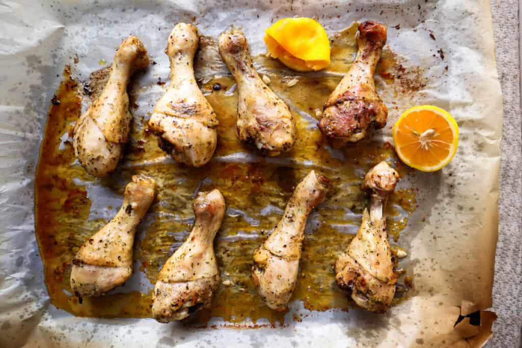 Eight baked chicken drumsticks on parchment paper with two halves of a lemon.