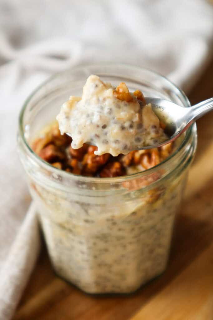 s\Spoonful of maple overnight oats with chopped pecans in a glass jar, which is on a wooden board.