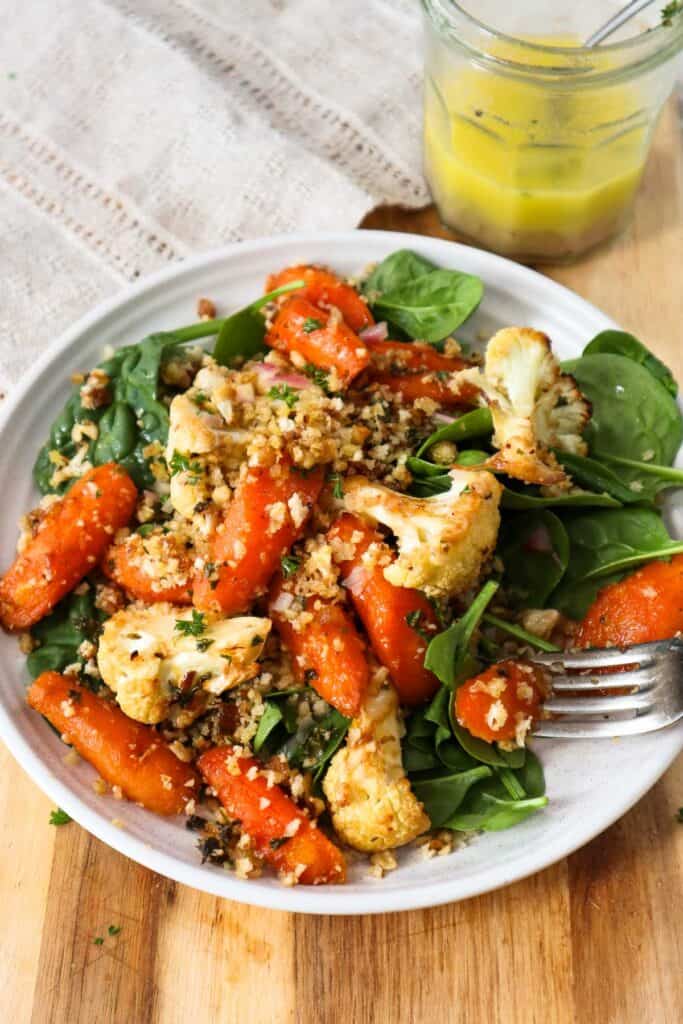 Roasted carrots and cauliflower on bed of baby spinach on a plate with salad dressing in a small jar.