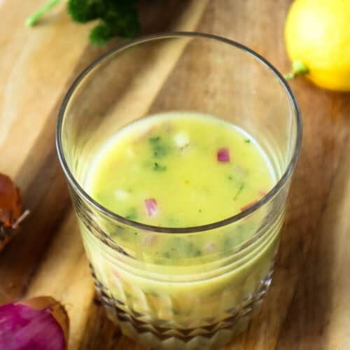 Lemon shallot vinaigrette in glass cup on wooden board with shallots and lemon and parsley.