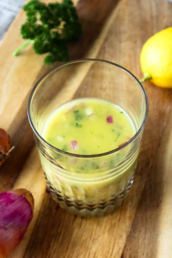 Lemon shallot vinaigrette in glass cup on wooden board with shallots and lemon and parsley.
