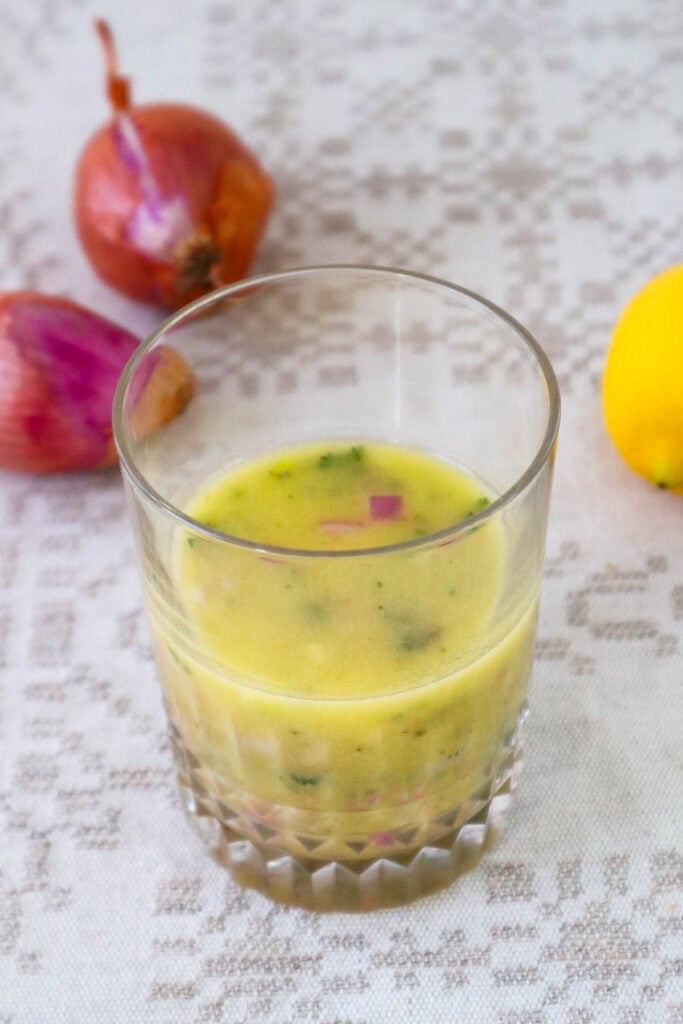 Overhead view of lemon shallot vinaigrette in glass cup on tablecloth.