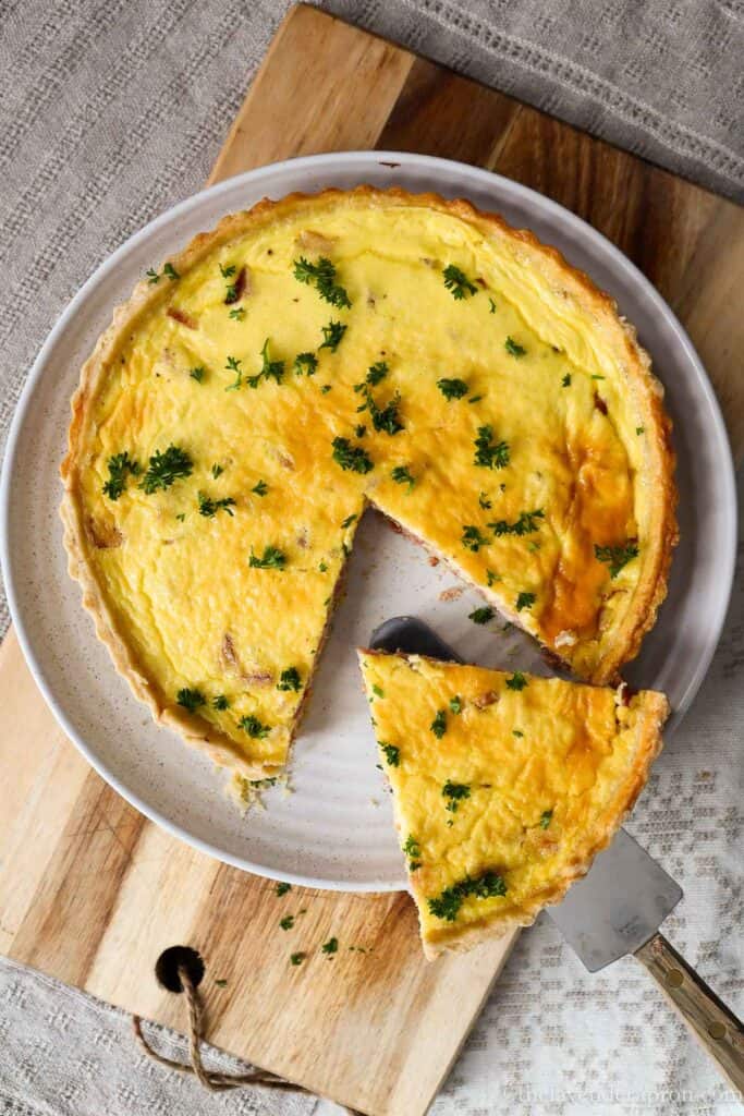 Slice of authentic French quiche Lorraine taken out of a whole quiche on a beige plate.