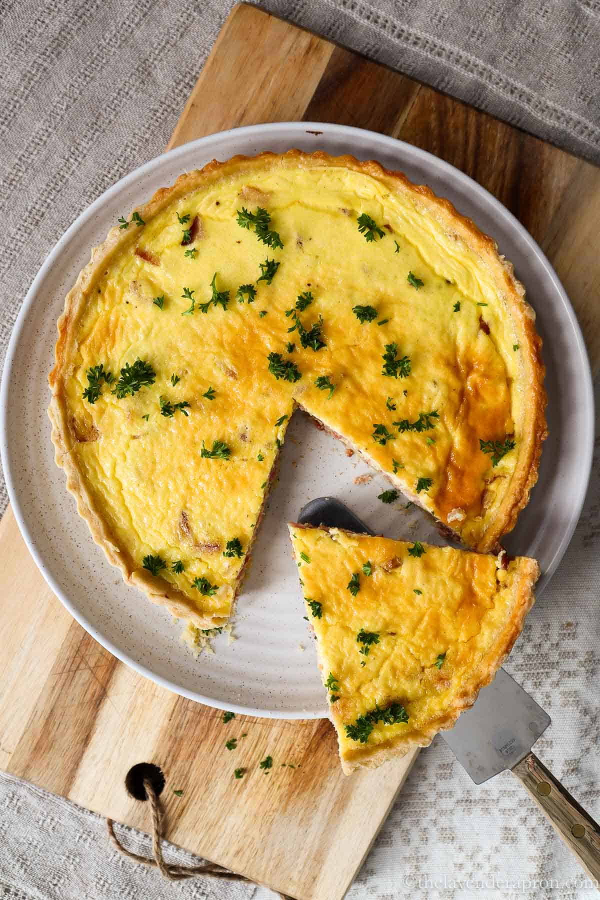 Slice of authentic French quiche Lorraine taken out of a whole quiche on a beige plate.