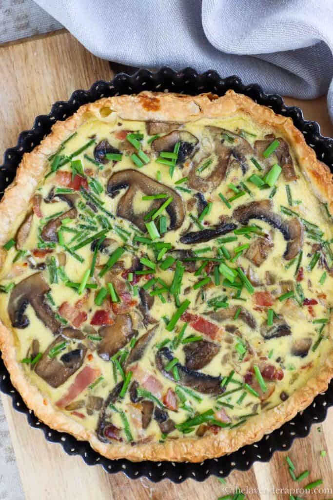 Quiche with mushrooms, bacon and chopped chives on top on wooden board, view from top.