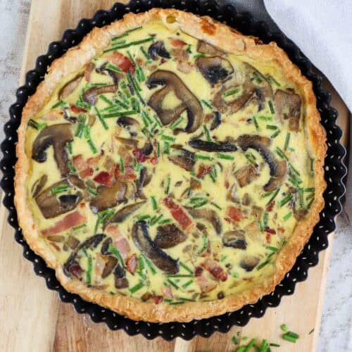 Whole bacon and mushroom quiche seen from top on wooden cutting board.