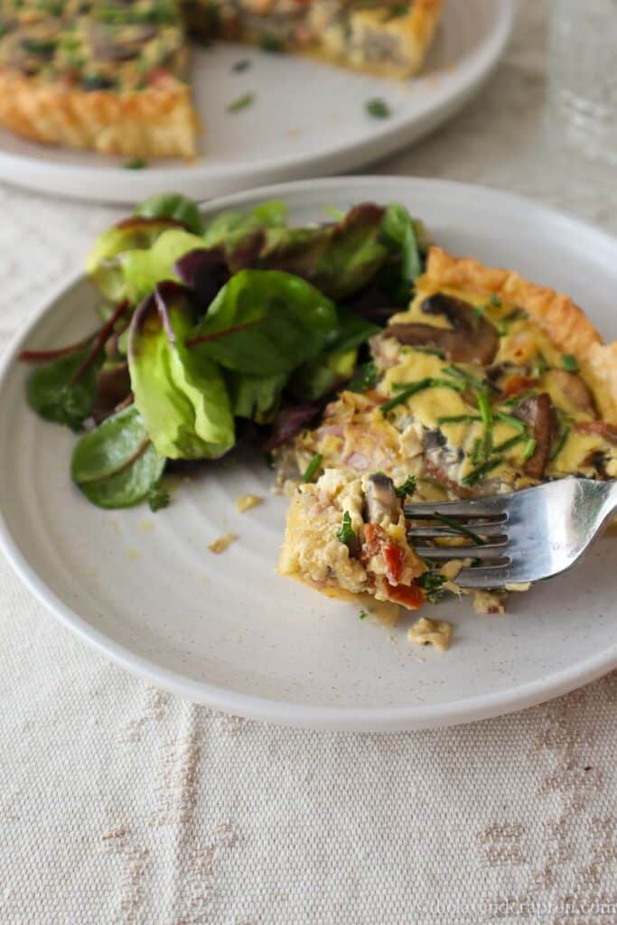Forkful of bacon mushroom quiche o beige plate next to a lettuce salad and large quiche in the background.