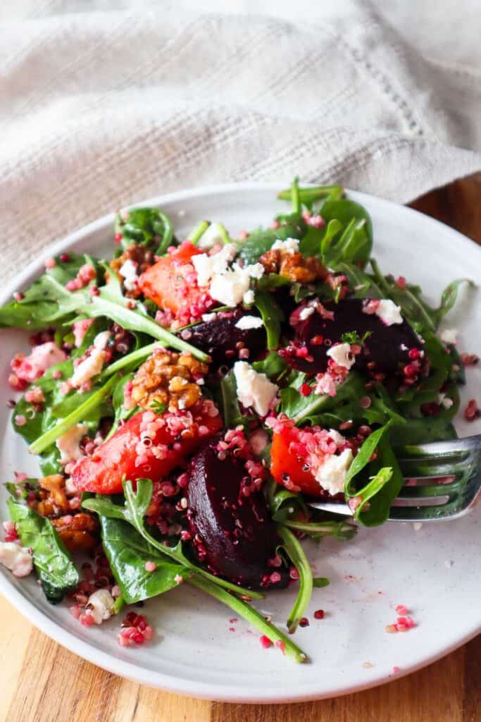 Forkful of fall harvest quinoa salad. There are carrots, beets, babay spinach, arugula, feta cheese, walnuts on beige plate.