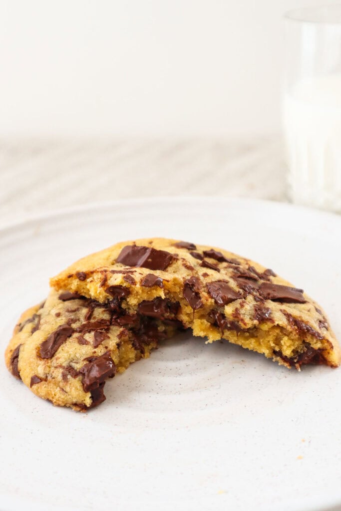 Single chocolate chip cookie broken in half on a plate with glass of milk in the background.