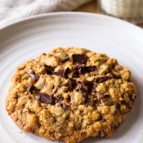 One oatmeal chocolate chip cookie on beige plate with glass of milk in background.