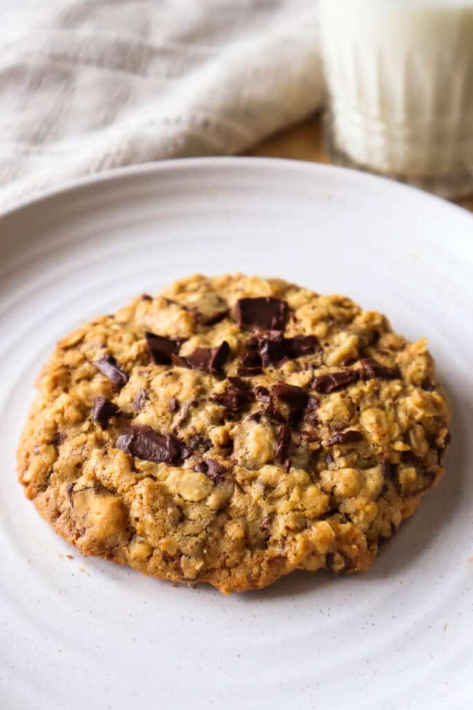 One oatmeal chocolate chip cookie on beige plate with glass of milk in background.