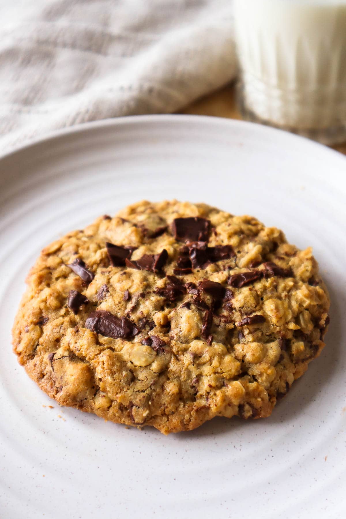One oatmeal chocolate chip cookie on beige plate with glass of milk in background.