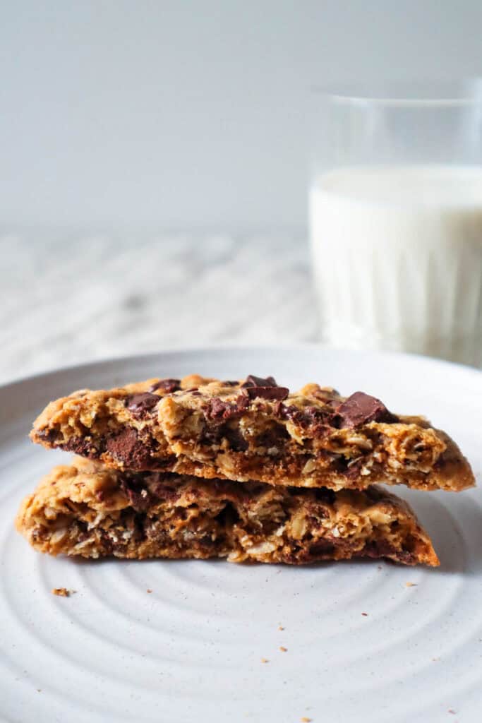 Single oatmeal chocolate chip cookie cut in half and one side is place on top of the other to show the chewy inside. Glass of milk in the background.