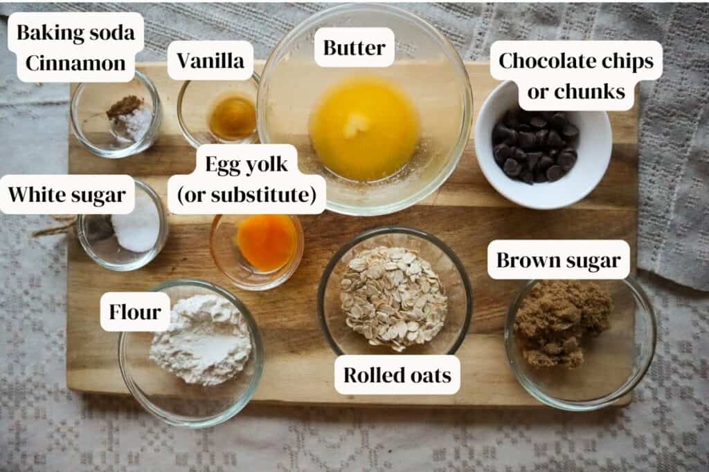 Chocolate chips, rolled oats, melted butter, vanilla, flour, broan and white sugar, egg yolk, baking soda and cinnamon in small glass bowls on a wooden cutting board.