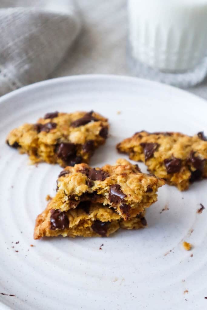 Pieces of oatmeal chocolate chip cookie on a beige plate with a glass of milk in the background.