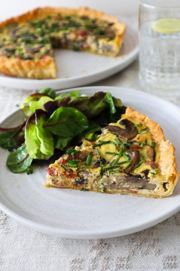 Slice of bacon mushroom quiche on plate with lettuce leaves and larger quiche in the background.