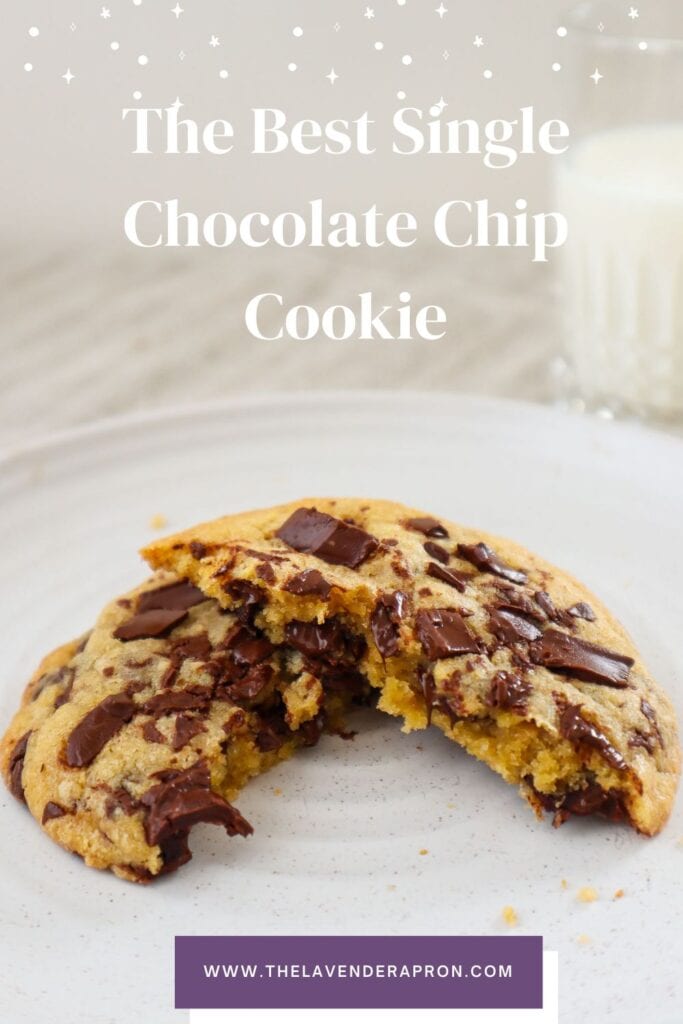 Single chocolate chip cookie cut in half on a beige plate with glass of milk in the background.