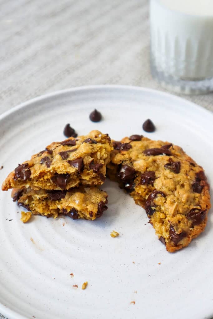 Large oatmeal chocolate chip cookie on a beige plate in three pieces with a glass of milk in the background.