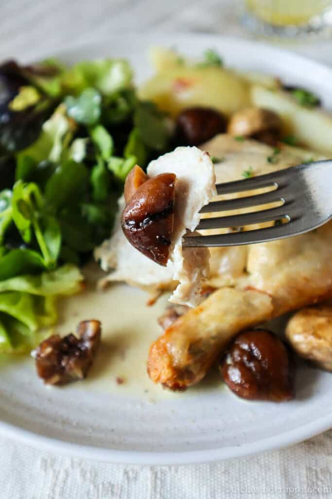 Close up of bite of chicken with piece of chestnut. There is a plate with green salad, chicken leg and chestnuts in the background.