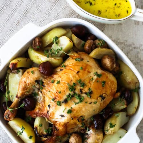 French roast chicken with chestnuts, mushrooms and potatoes with cider sauce on the side in a pouring dish.