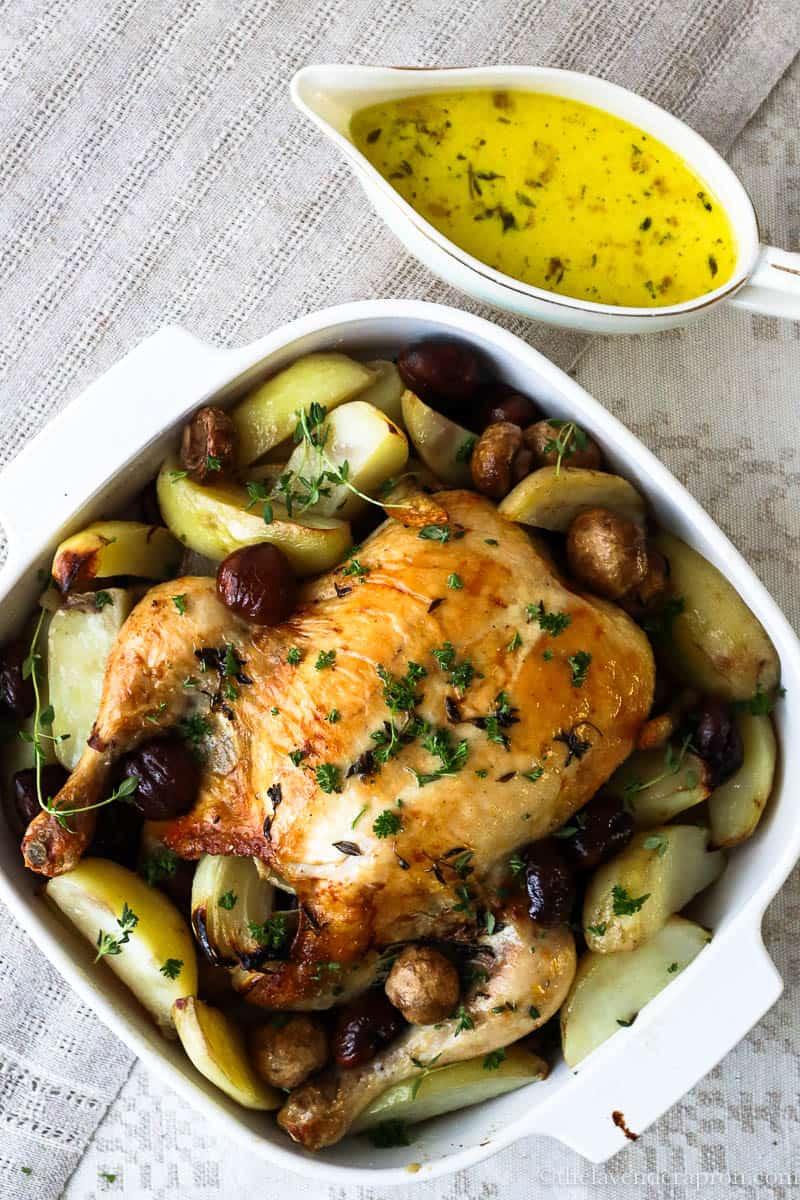 French roast chicken with chestnuts, mushrooms and potatoes with cider sauce on the side in a pouring dish.