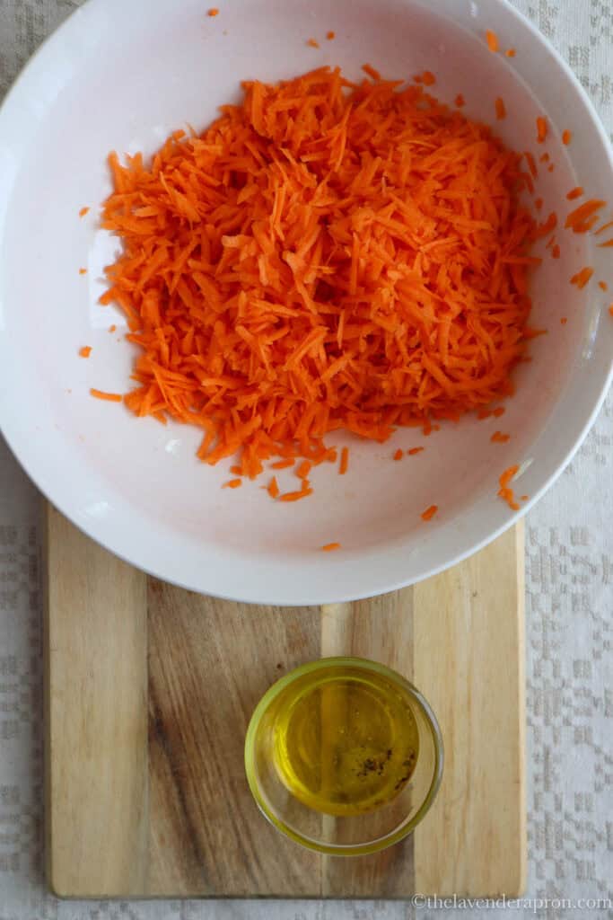 Grating carrots in a white bowl and olive oil and lemon juice in small clear bowl on wooden board.