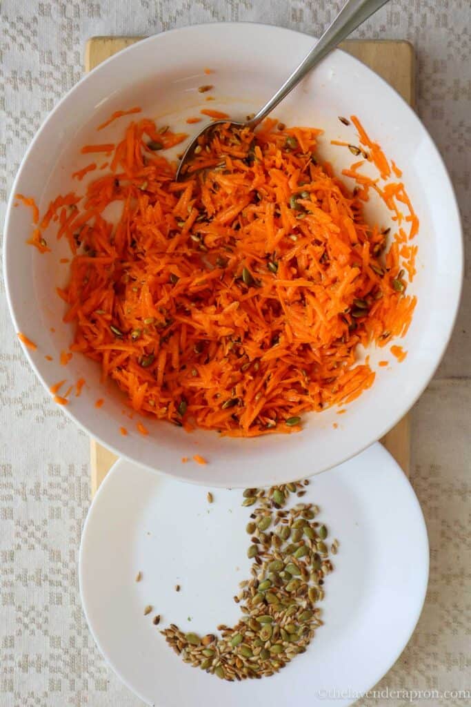 Mixing the grated carrots with dressing and pumpkin seeds and sunflower seeds in a white bowl. There are some leftover seeds on white plate.