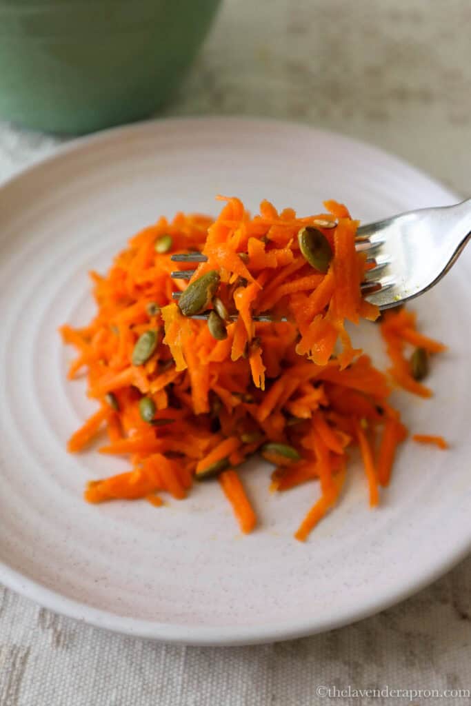 Fork of French carrot salad with pumpkin seeds and sunflower seeds on beige plate.
