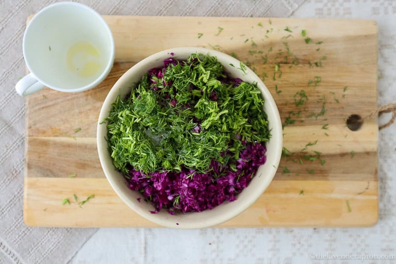 Finely sliced red cabbage in bowl topped with chopped dill and lemon dressing on a wooden board.
