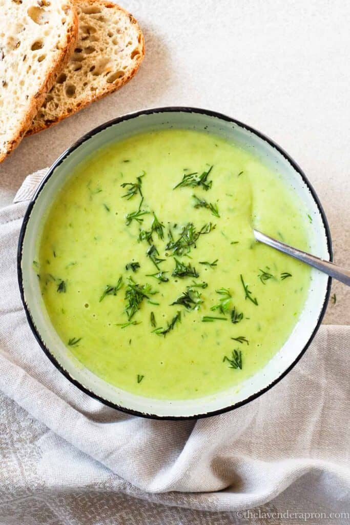 Asparagus and dill soup in a bowl with spoon and with two slices of grain bread next to it.