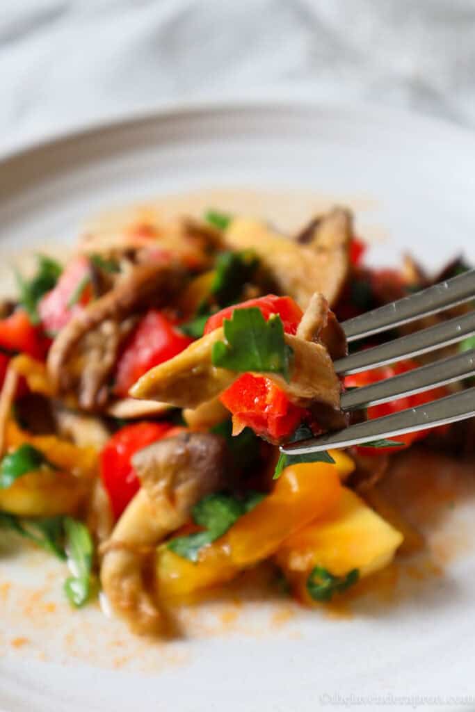 Close-up of a fork with mushroom and tomato and a plate of mushrooms and diced tomatoes in the background.