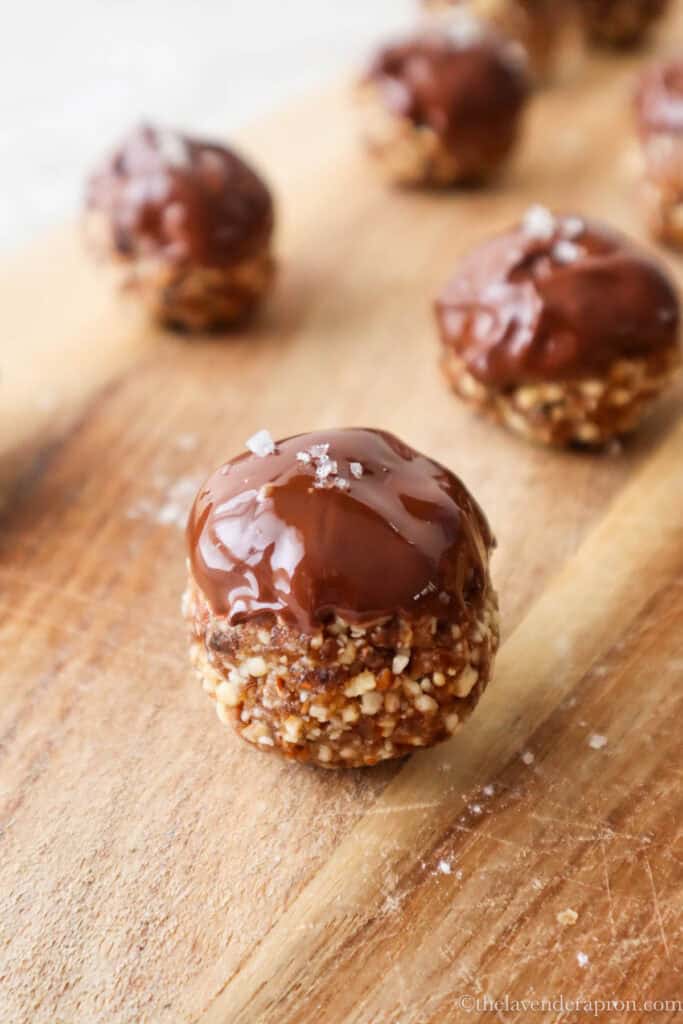 Date cashew almond balls topped with dark chocolate and a pinch of sea salt on a wooden chopping board.