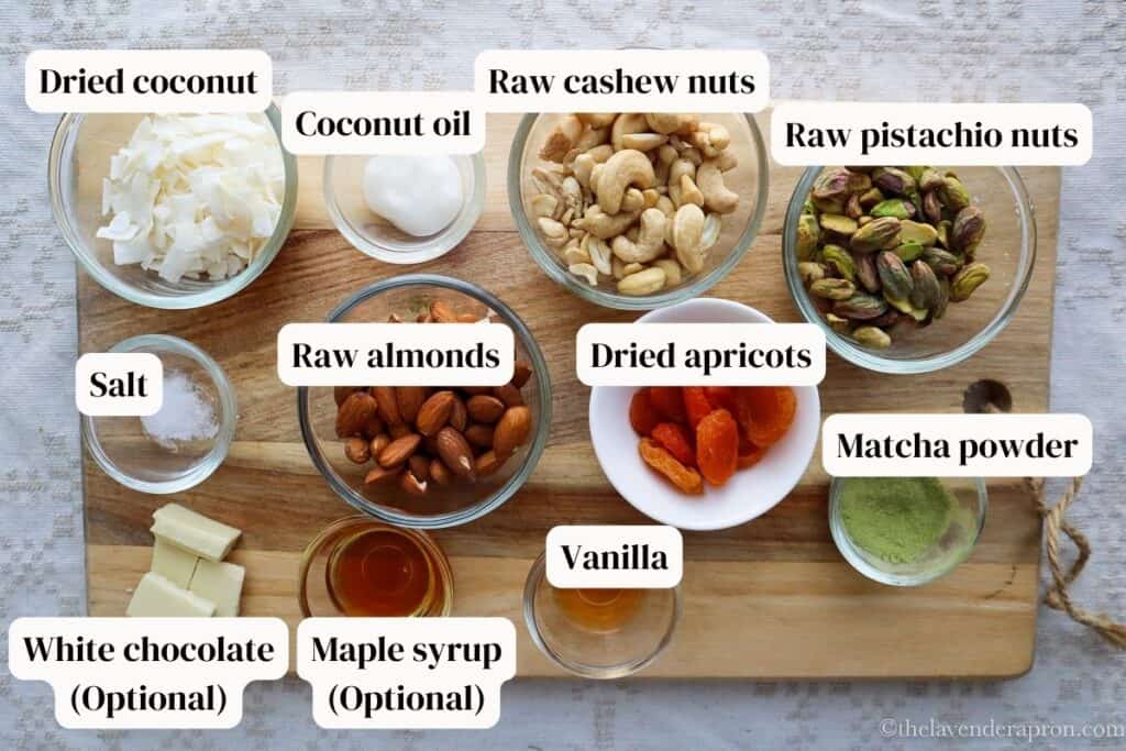 Cashews, almonds, pistachios, coconut, apricots, coconut oil, vanilla, salt, Matcha, and maple syrup on a wooden board. They are the ingredients to make pistachio matcha energy balls.