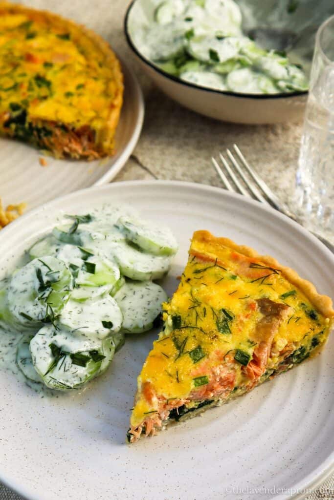 A slice of spinach salmon quiche served in a beige plate with a sliced cucumber creamy salad.