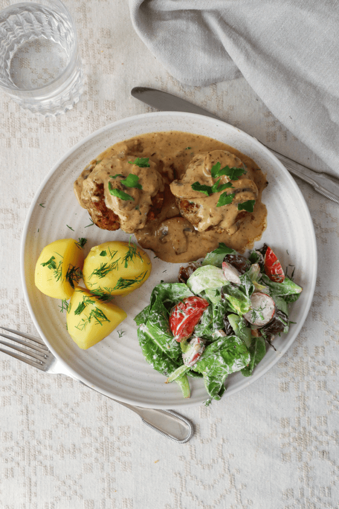 Overhead view of two kotletes covered in mushroom sauce, boiled potatoes with dill and a green salad with tomatoes.
