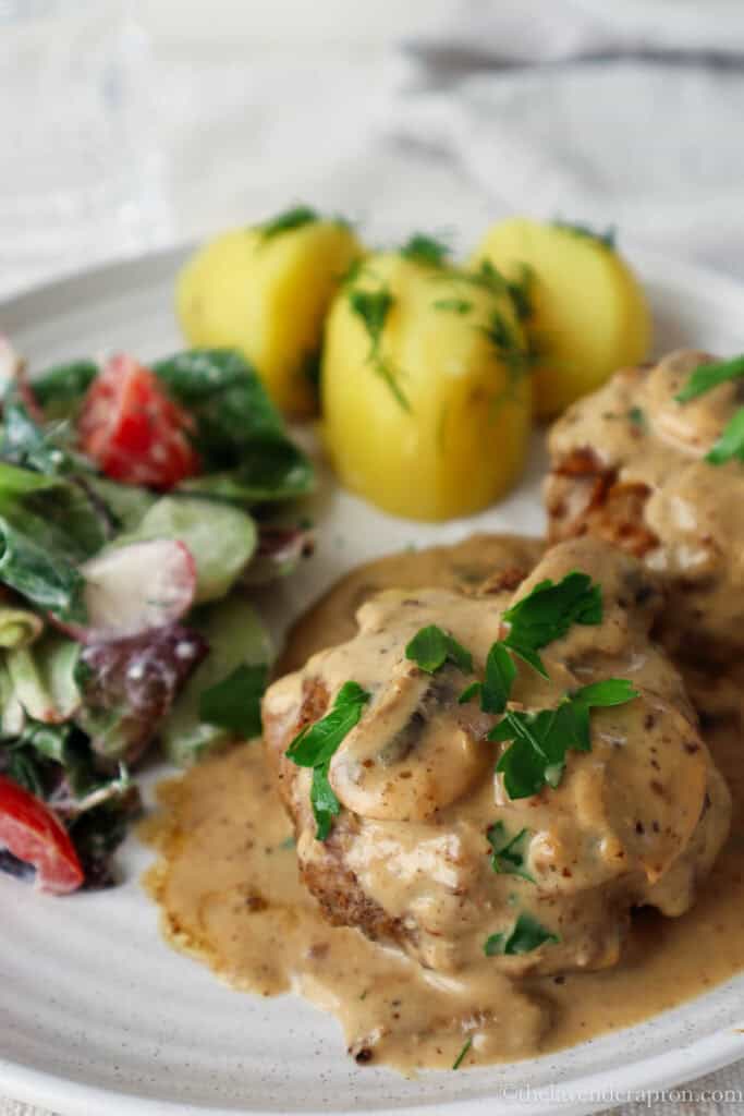 Latvian kotlete covered with lots of mushroom cream sauce on a plate with salad and potato.