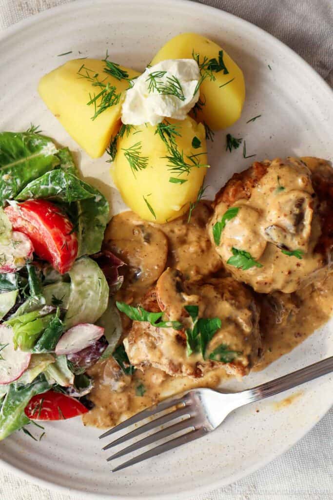 Latvian kotletes covered in cream mushroom sauce served with potatoes, sour cream and salad.