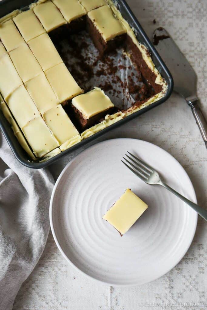Belgian white chocolate ganache on top of Belgian chocolate cake in square pan with a small slice on a beige plate.