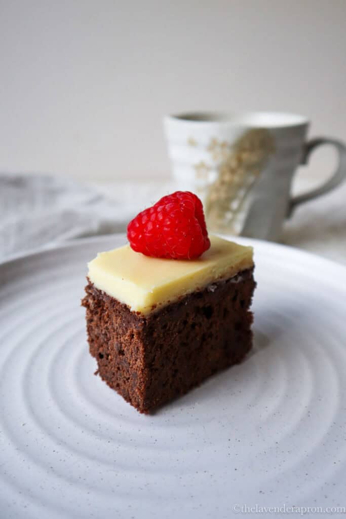 Belgian chocolate cake with white chocolate ganache and a raspberry served on a beige plate with coffee cup in background.