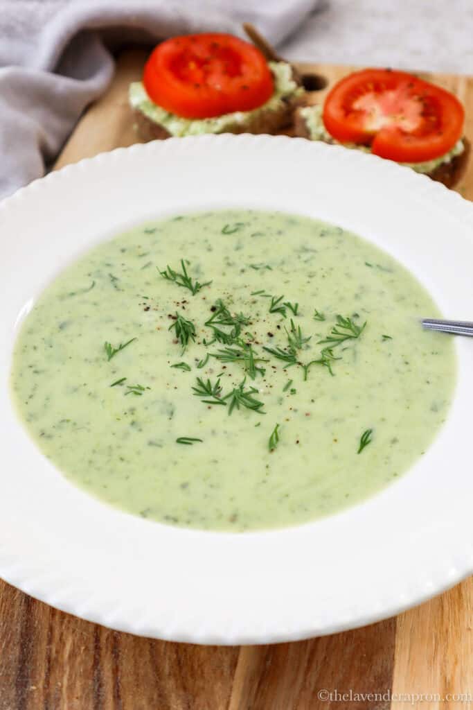 Creamy zucchini dill soup served with cottage cheese and tomato sandwiches on a wooden board.