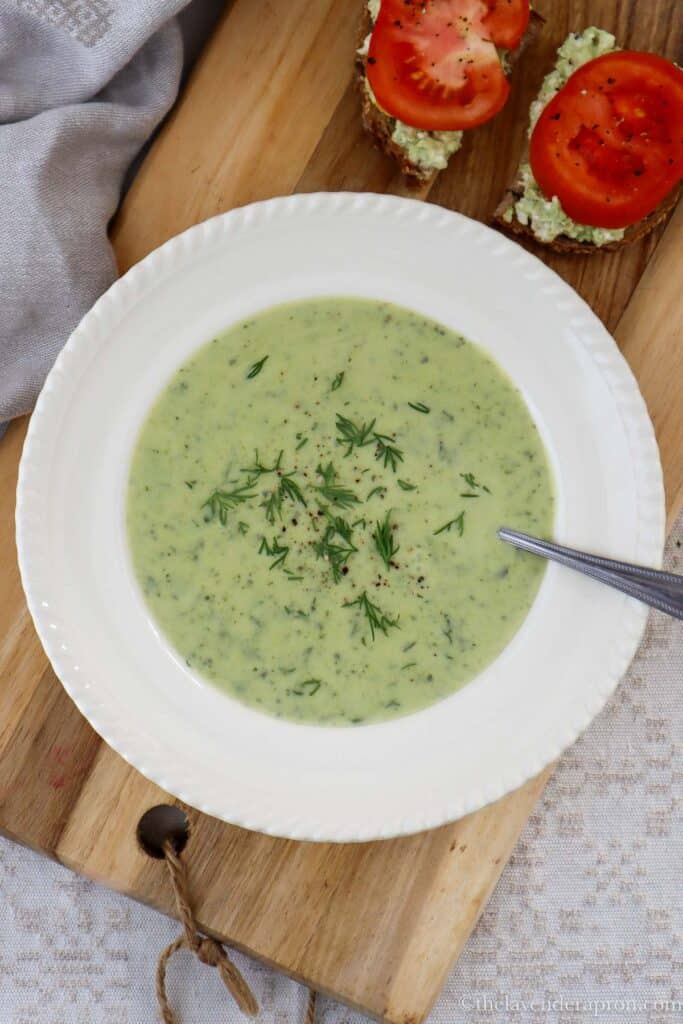 Creamy zucchini dill soup served with cottage cheese and tomato sandwiches on a wooden board.