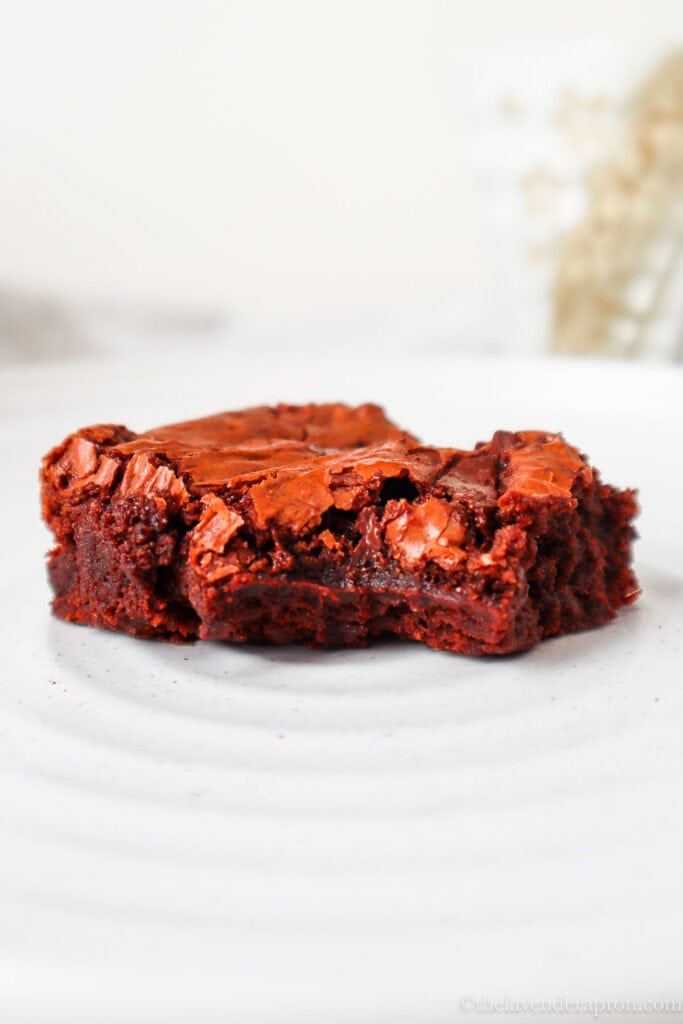 Moist chocolate brownie with crackkly top on a beige plate with coffee cup in the background.