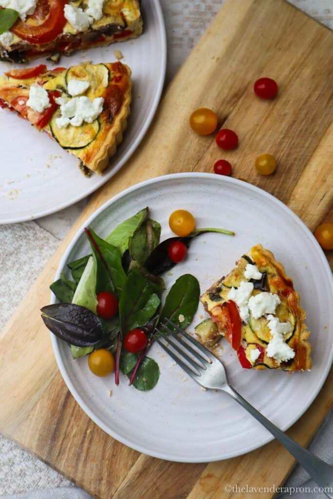 Quiche Provencale served with a green salad and small tomatoes on a beige plate.