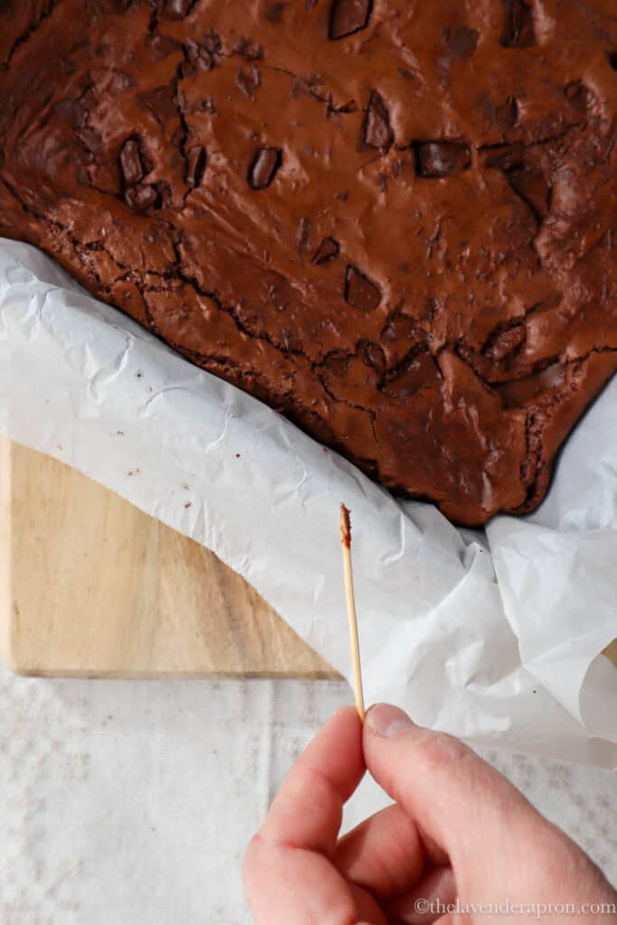 Toothpick showing batter on it next to baked brownies in square tin.